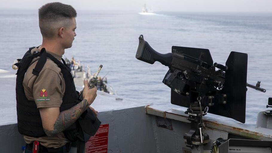 A U.S. sailor in a tan shirt and life vest stands watch at a deck gun, radio in hand, with another ship on the horizon